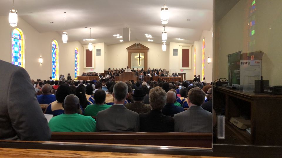 Soundcheck with musicians and choir on the platform under the lit cross, white loudspeakers hanging from the ceiling, empty pews in the foreground.