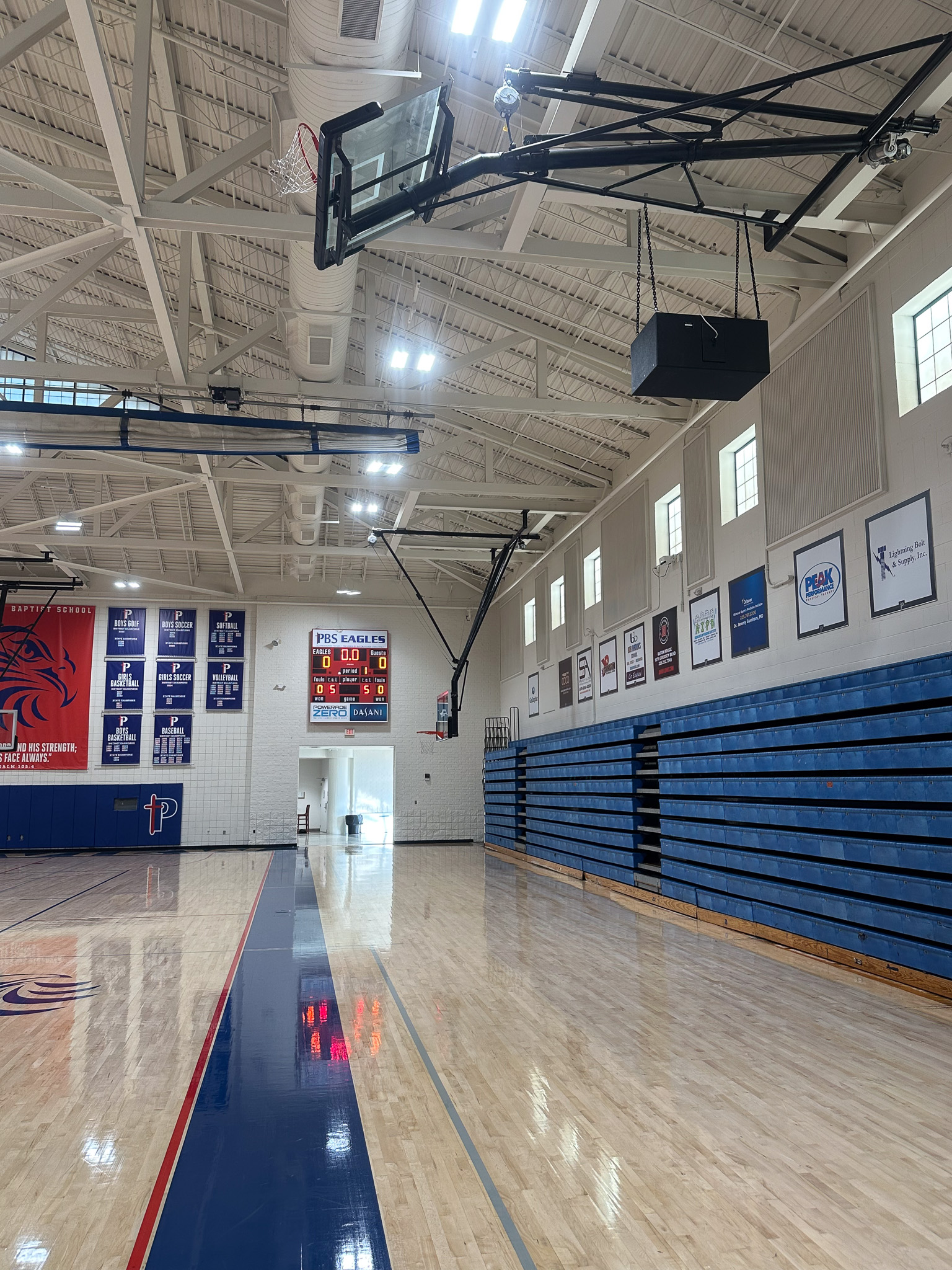 Parkview Eagles gym from court level — folded basketball hoop and Blaze 528 loudspeaker hung overhead from chains in the foreground, championship banners stacked along the wall on the left, PBS Eagles scoreboard at the back, blue padded bleachers along the right wall.