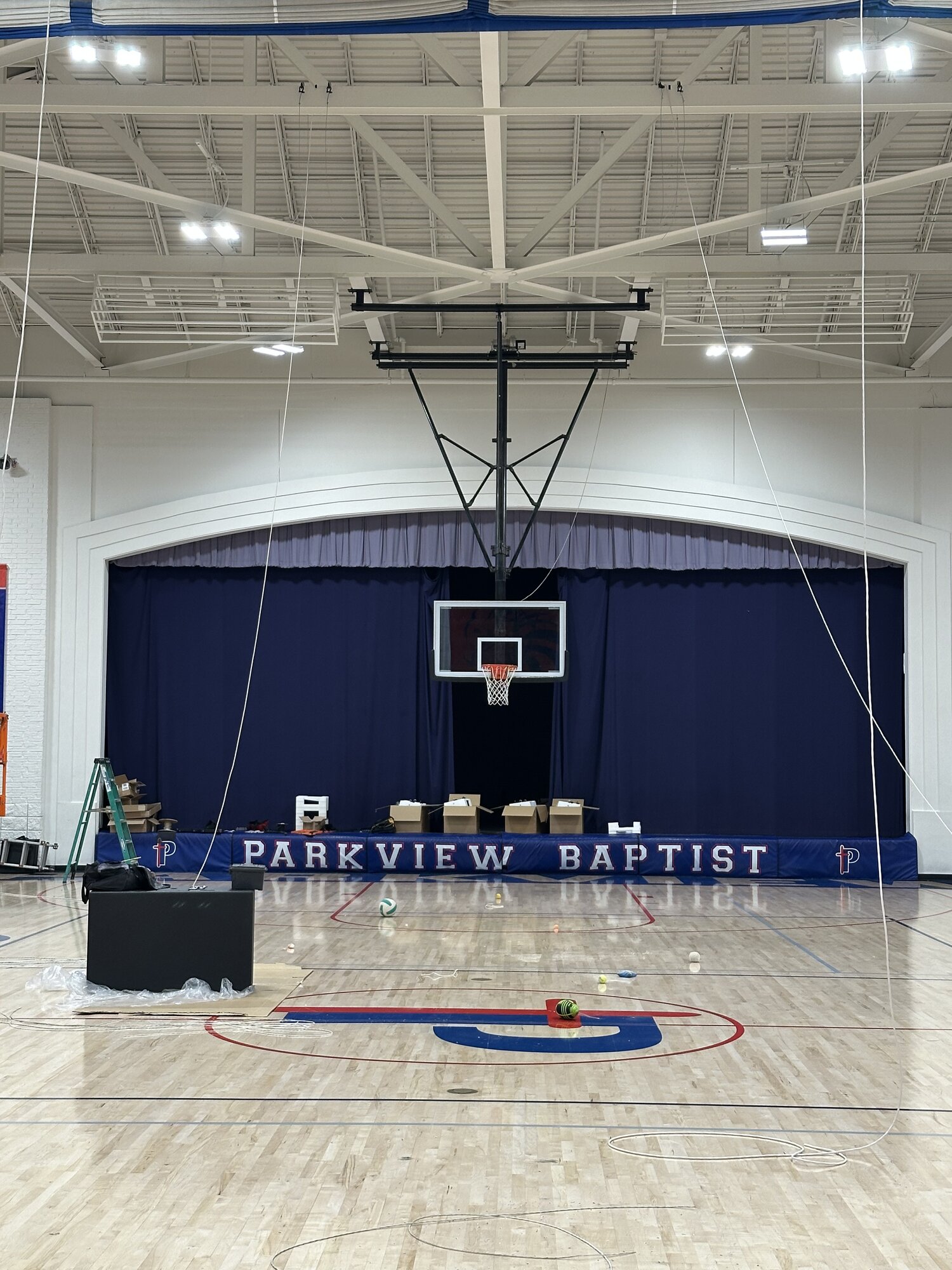 Install in progress — scissor lift extended to ceiling height with a technician mounting a Blaze 528 loudspeaker from the structure above the bleachers.