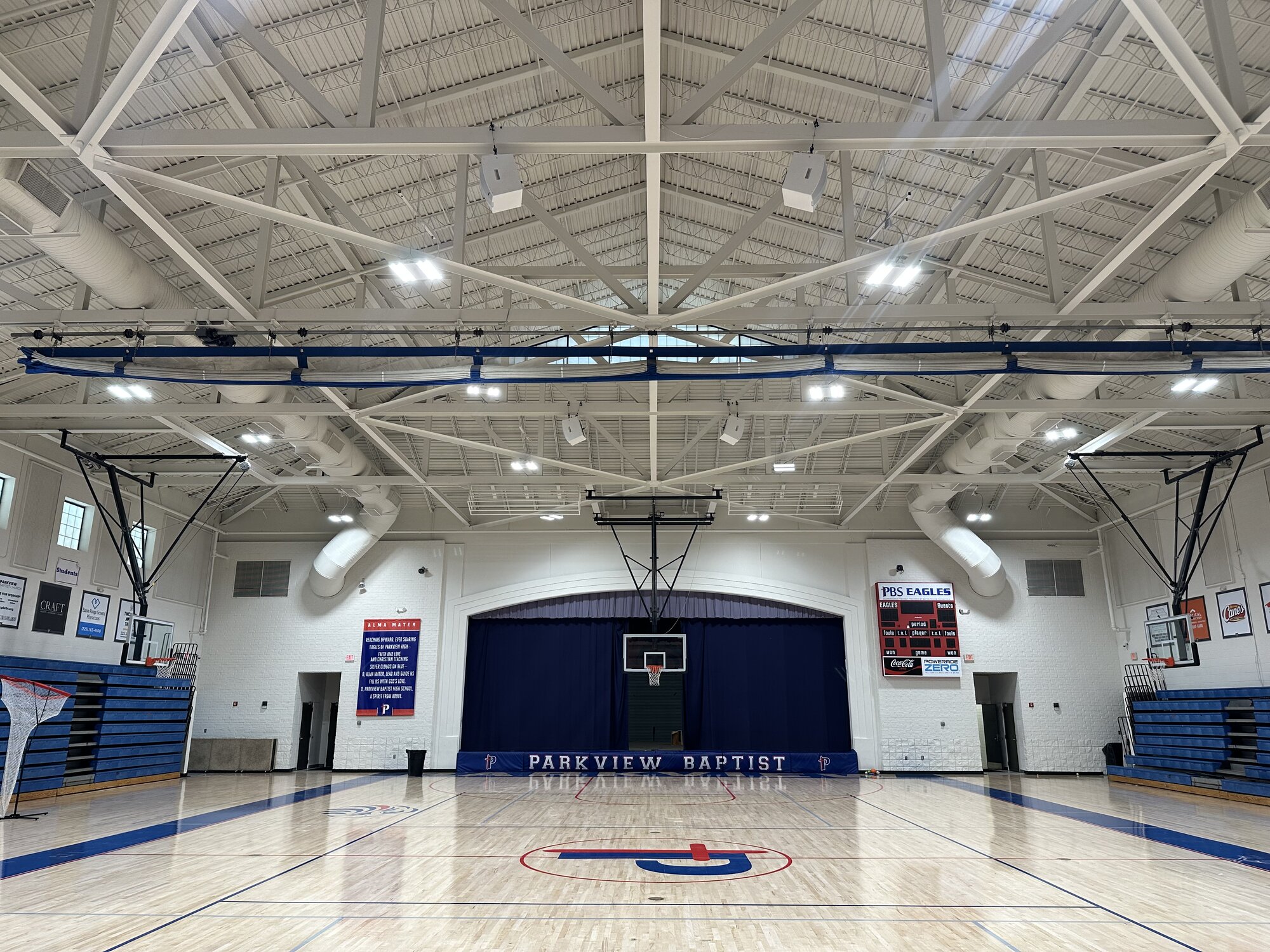 Parkview Baptist School Eagles gym — wide court-level view showing the Eagles logo at center court, Blaze 528 loudspeakers hung from the structural ceiling, scoreboard and championship banners, polished wood floor.